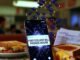 Cracked iPhone with "Free College Bill" notification on greasy table with grilled cheese and overdue bills, 90s VHS style, pixelated graduation caps falling.
